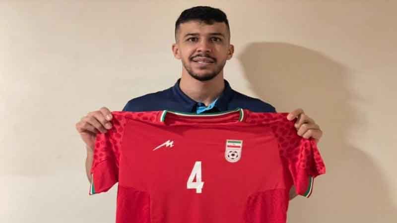 بازیکنان تیم ملی فوتسال ایران با لباس‌های سرخ‌پوش در مقابل تیم امارات با لباس سفید، در زمین مسابقه صف‌آرایی کرده‌اند. پرچم‌های ایران و امارات در بالا دیده می‌شوند و ساعت مسابقه ۱۱:۳۰ را نشان می‌دهد.